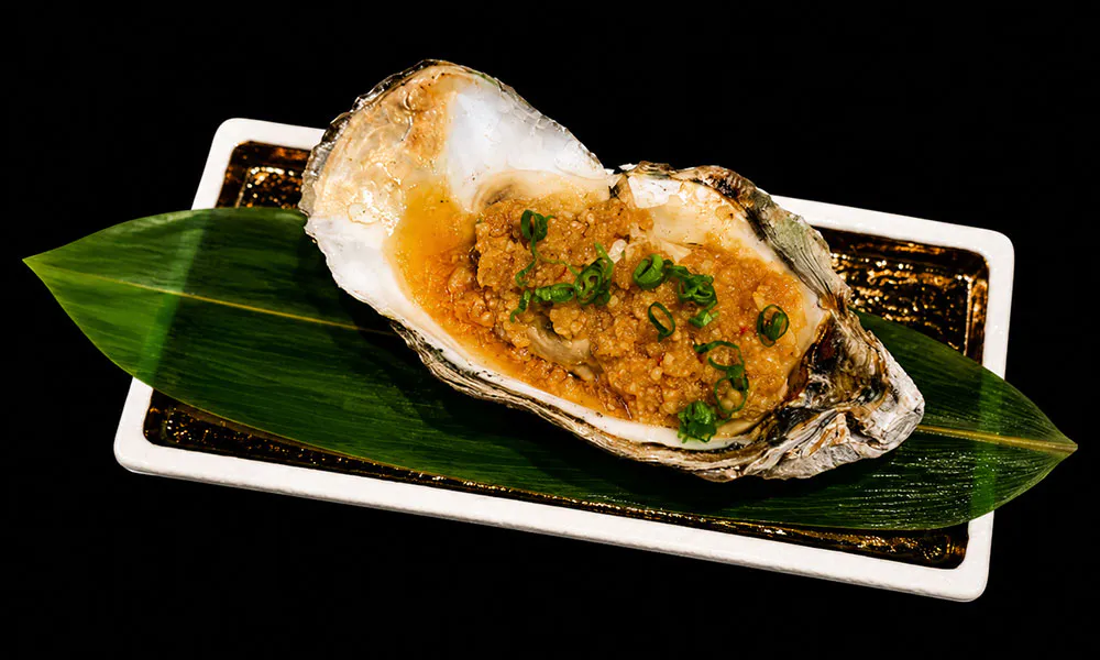 Garlic baked oyster topped with minced garlic and green scallions on bamboo leaf rectangular plate at Sat Asian Seafood Restaurant &Bar, a Yakitori Restaurant in Flushing