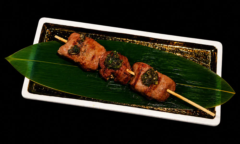 Grilled beef skewer topped with herbs laid on bamboo leaf decorated rectangular dish at Sat Asian Seafood Restaurant &Bar, a Yakitori Restaurant in Flushing